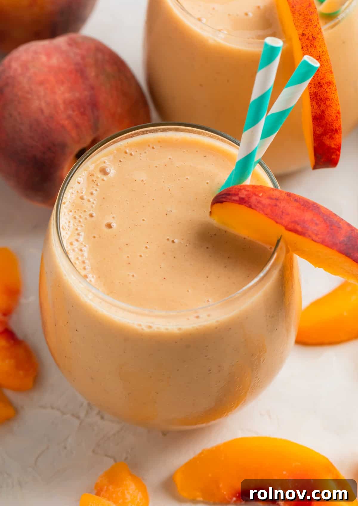 A tall glass filled with a creamy banana peach smoothie, garnished with two straws and a fresh peach slice on the rim. The smoothie showcases its smooth texture and inviting peachy hue.