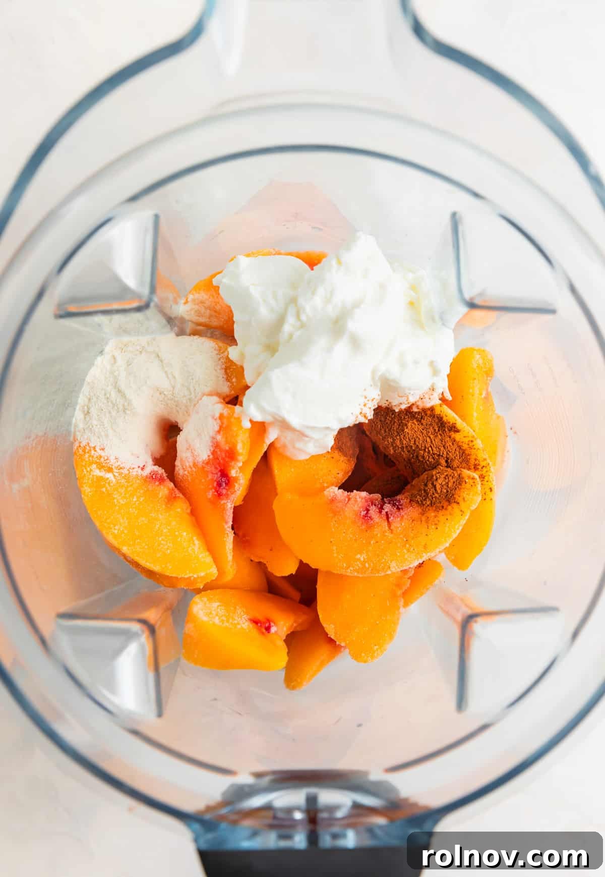 All the smoothie ingredients, including sliced peaches, frozen banana, milk, and yogurt, are visible inside a blender pitcher, ready to be blended.