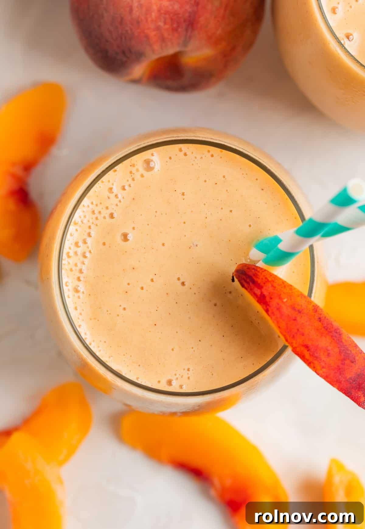 A vibrant peach and banana smoothie served in a clear glass cup, garnished with a fresh peach slice on the rim and a stylish teal and white striped straw. The focus is on the smoothie's inviting color and smooth texture.