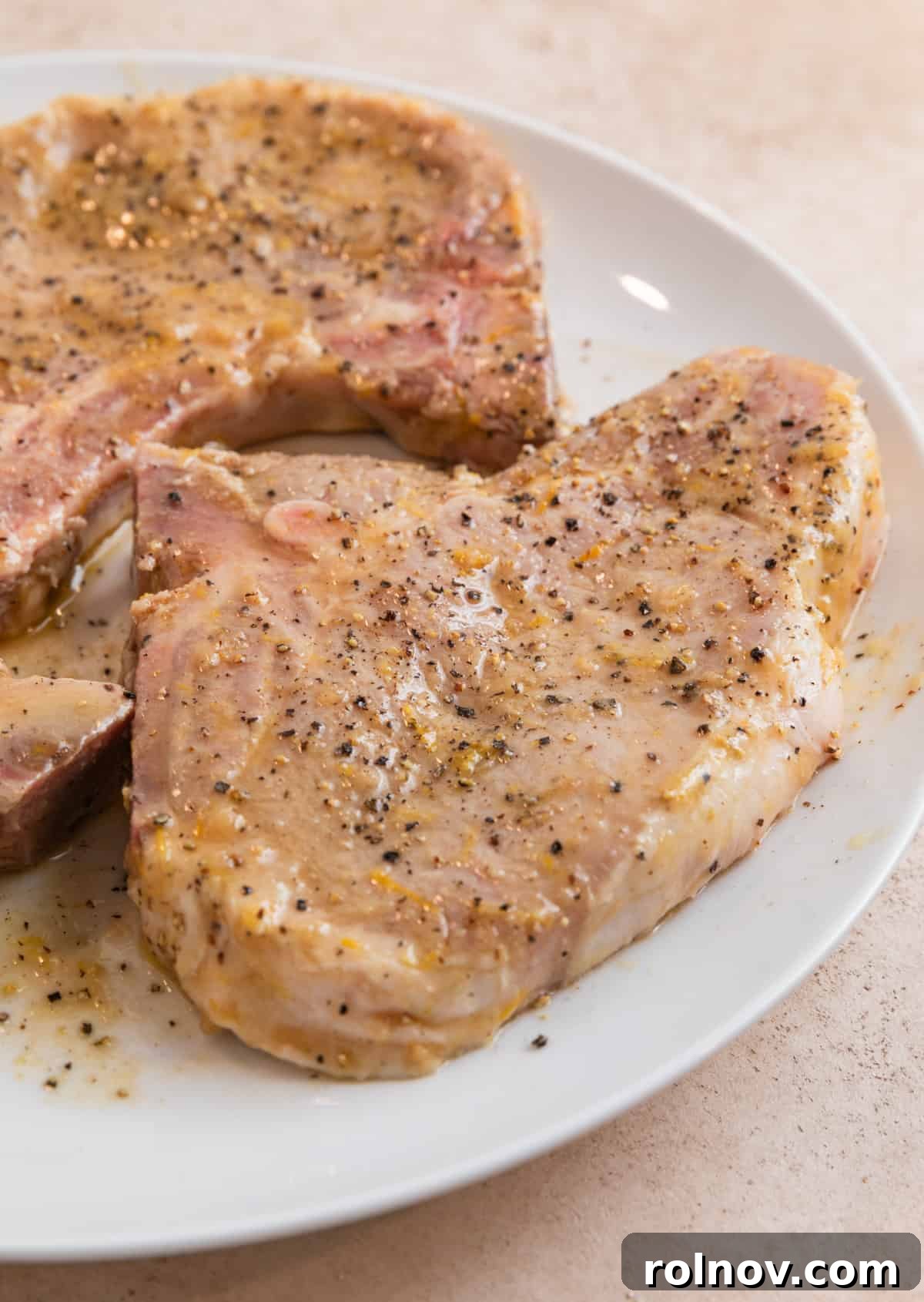 Zesty Lemon Pepper Pork Chops 6 Marinated lemon pepper pork chops resting on a white plate, seasoned with additional cracked pepper, ready for searing.