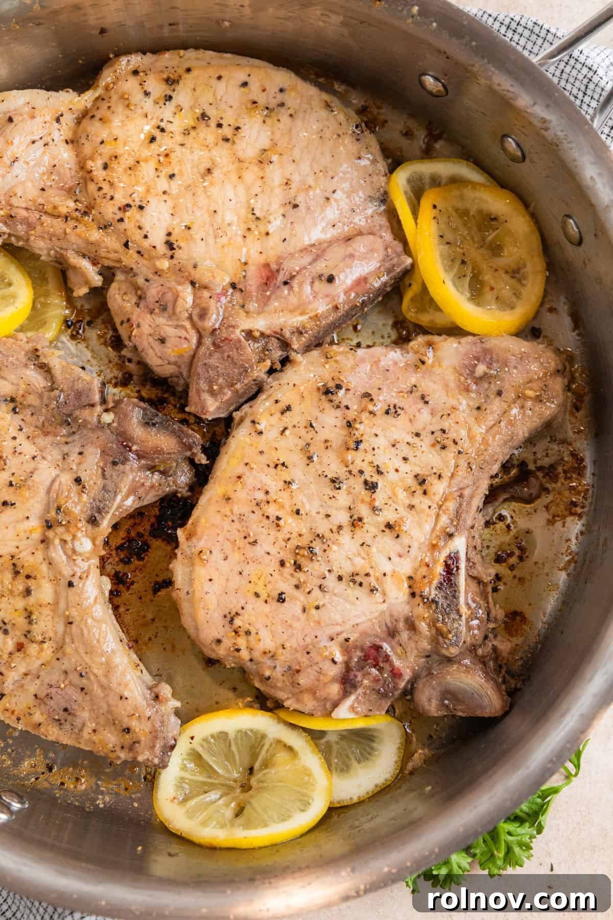 Zesty Lemon Pepper Pork Chops 7 Three lemon pepper pork chops searing to golden perfection in a stainless steel skillet, with thin lemon slices adding flavor.