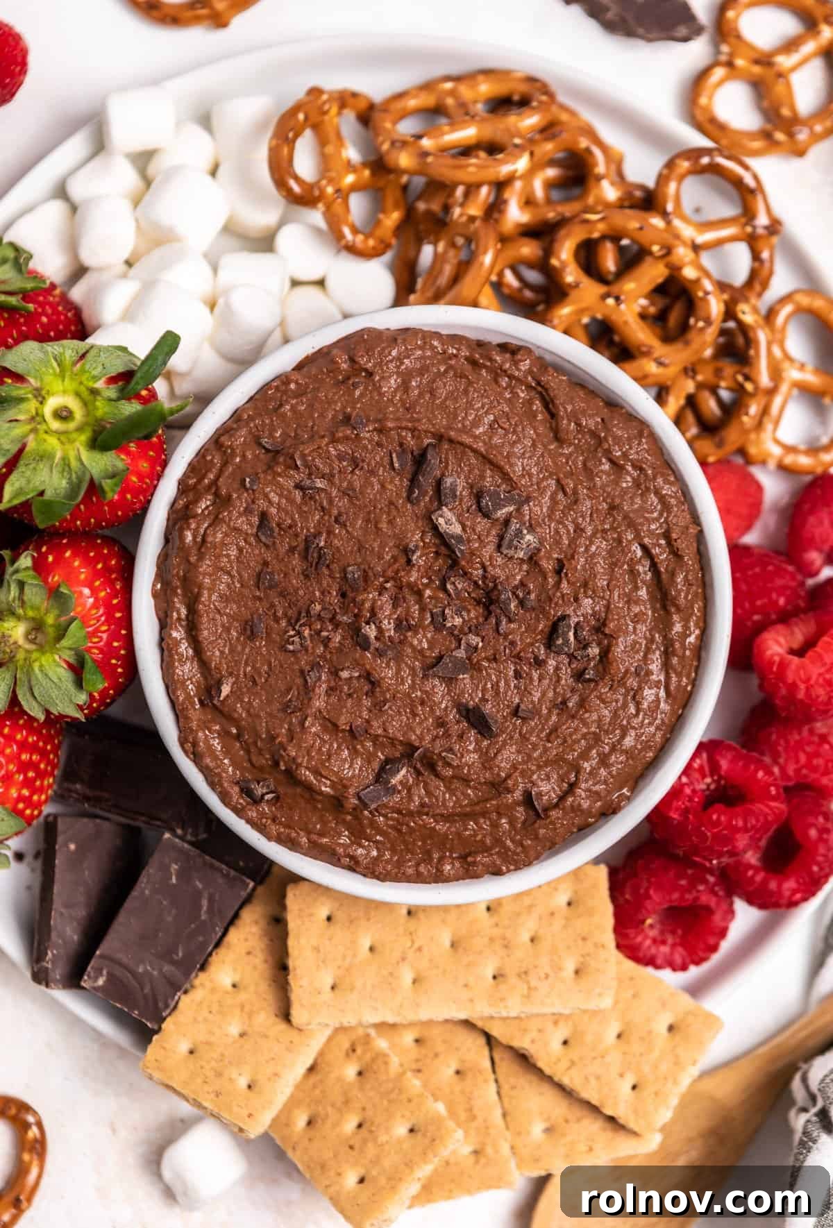 Bowl of chocolate brownie batter hummus with chopped chocolate on top on plate with marshmallows, pretzels and berries.