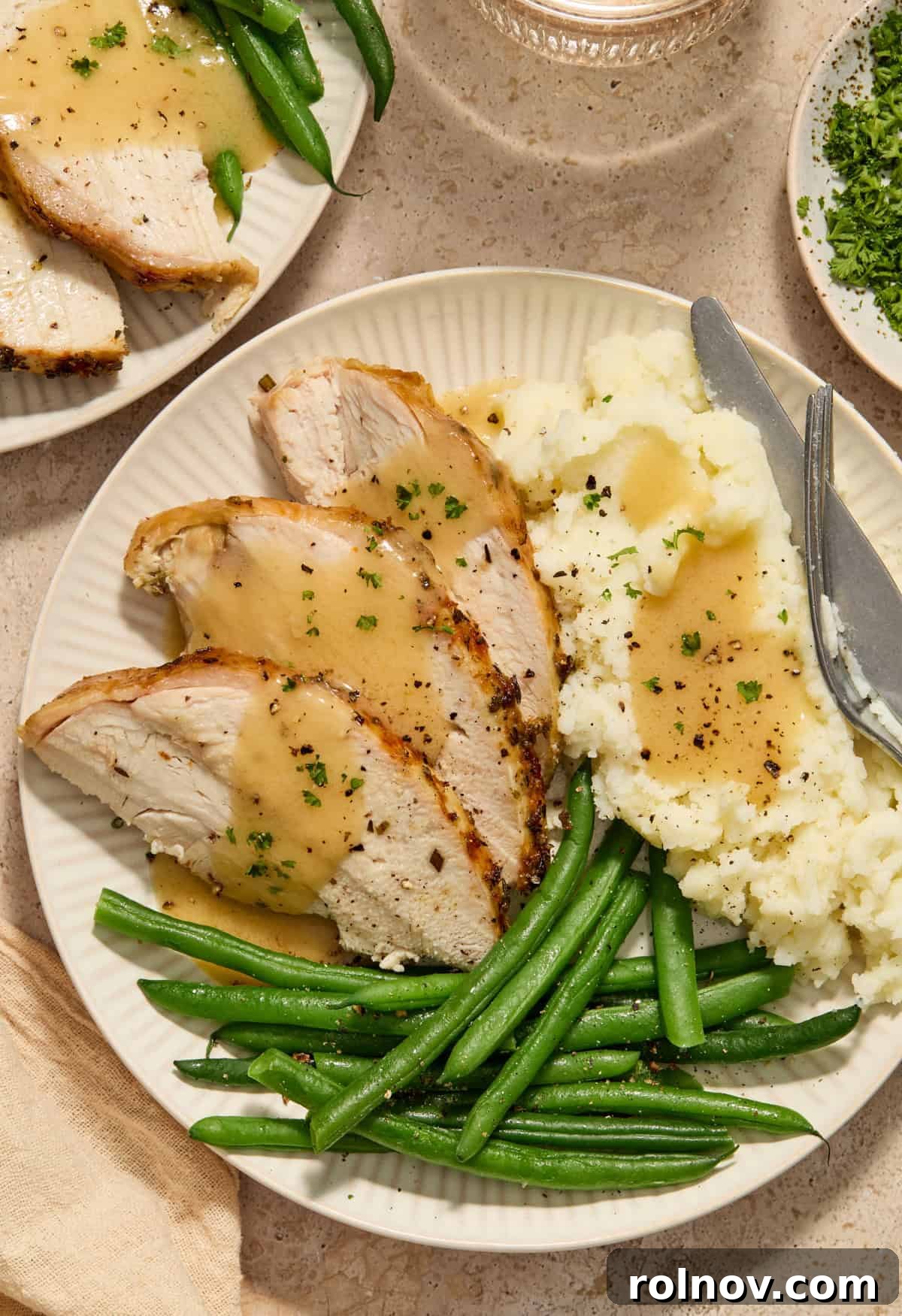 Slices of turkey with gravy, cooked green beans and mashed potatoes on plate with fork and knife.
