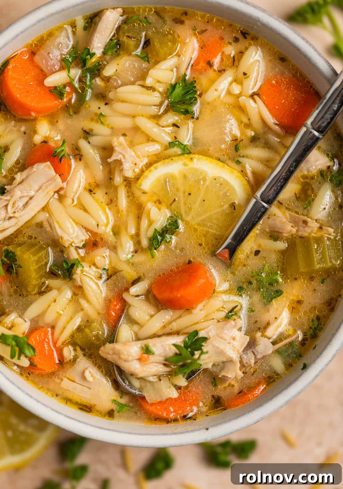 Zesty Lemon Chicken Orzo Soup 2 A steaming bowl of Chicken Lemon Orzo Soup, garnished with fresh lemon slices and chopped parsley, with a spoon gently scooping a hearty portion.