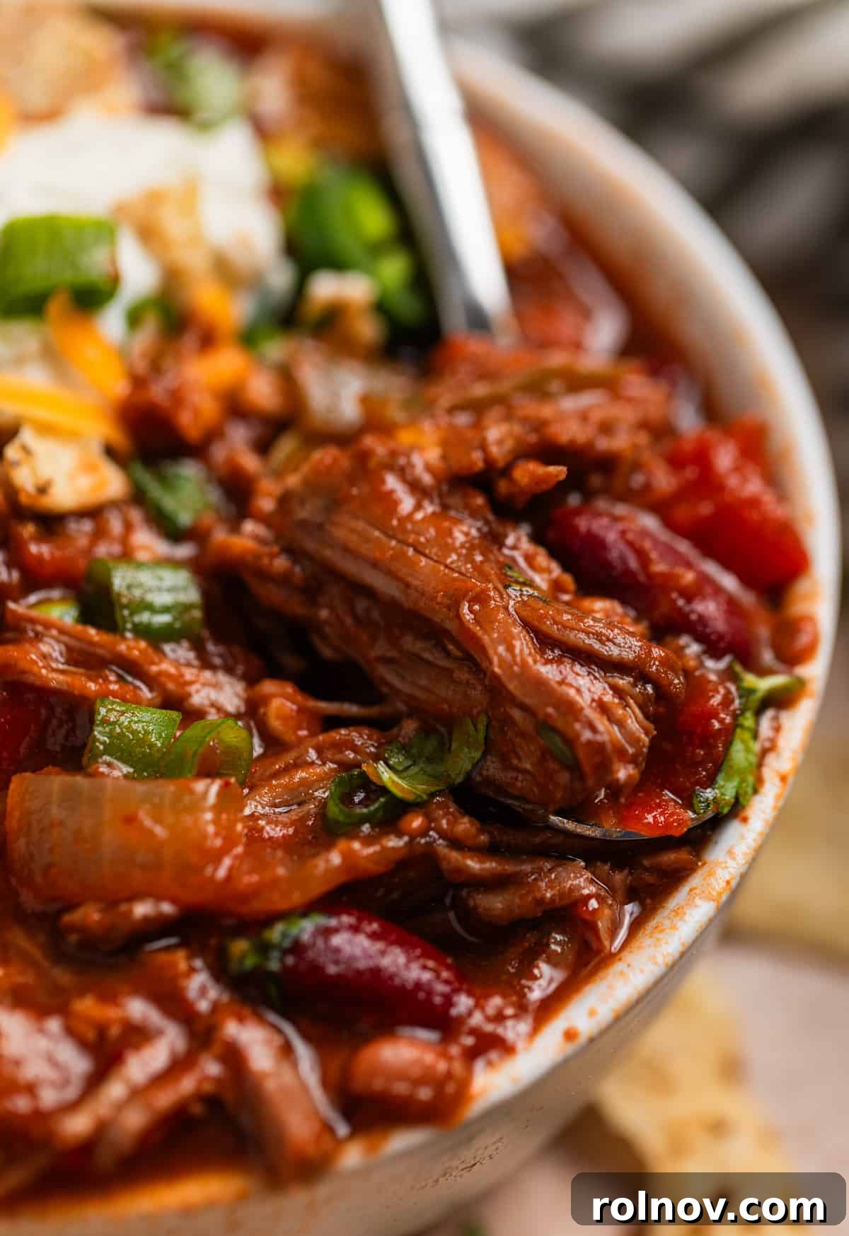 Slow Cooker Pulled Beef Chili 2 Bowl of tender slow cooker shredded beef chili with a spoon scooping into its rich depths, showcasing the hearty beef, beans, and vibrant spices.