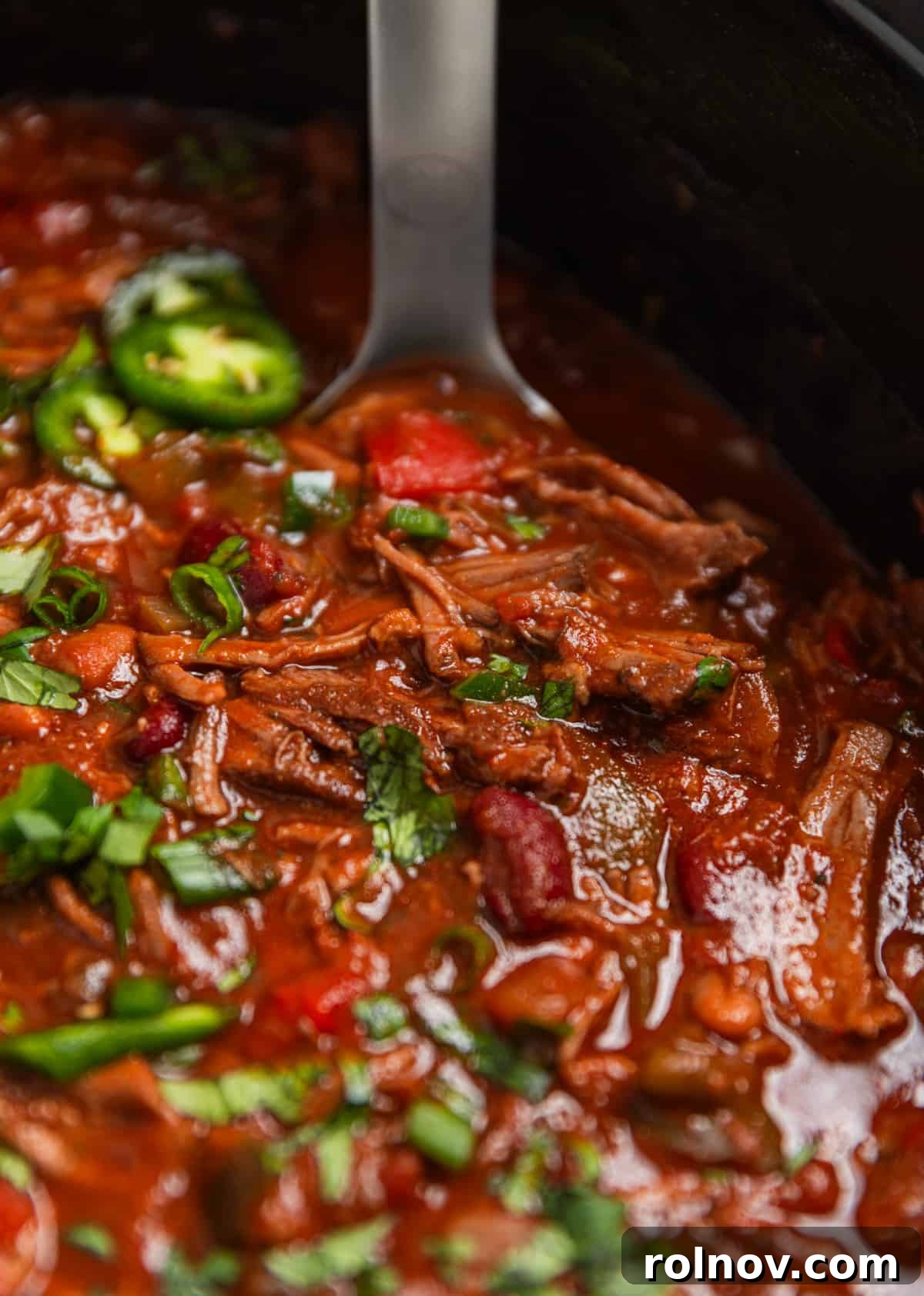 Slow Cooker Pulled Beef Chili 11 Close-up of slow cooker shredded beef chili in the crockpot, garnished with fresh cilantro and sliced jalapeño, with a ladle ready to serve.