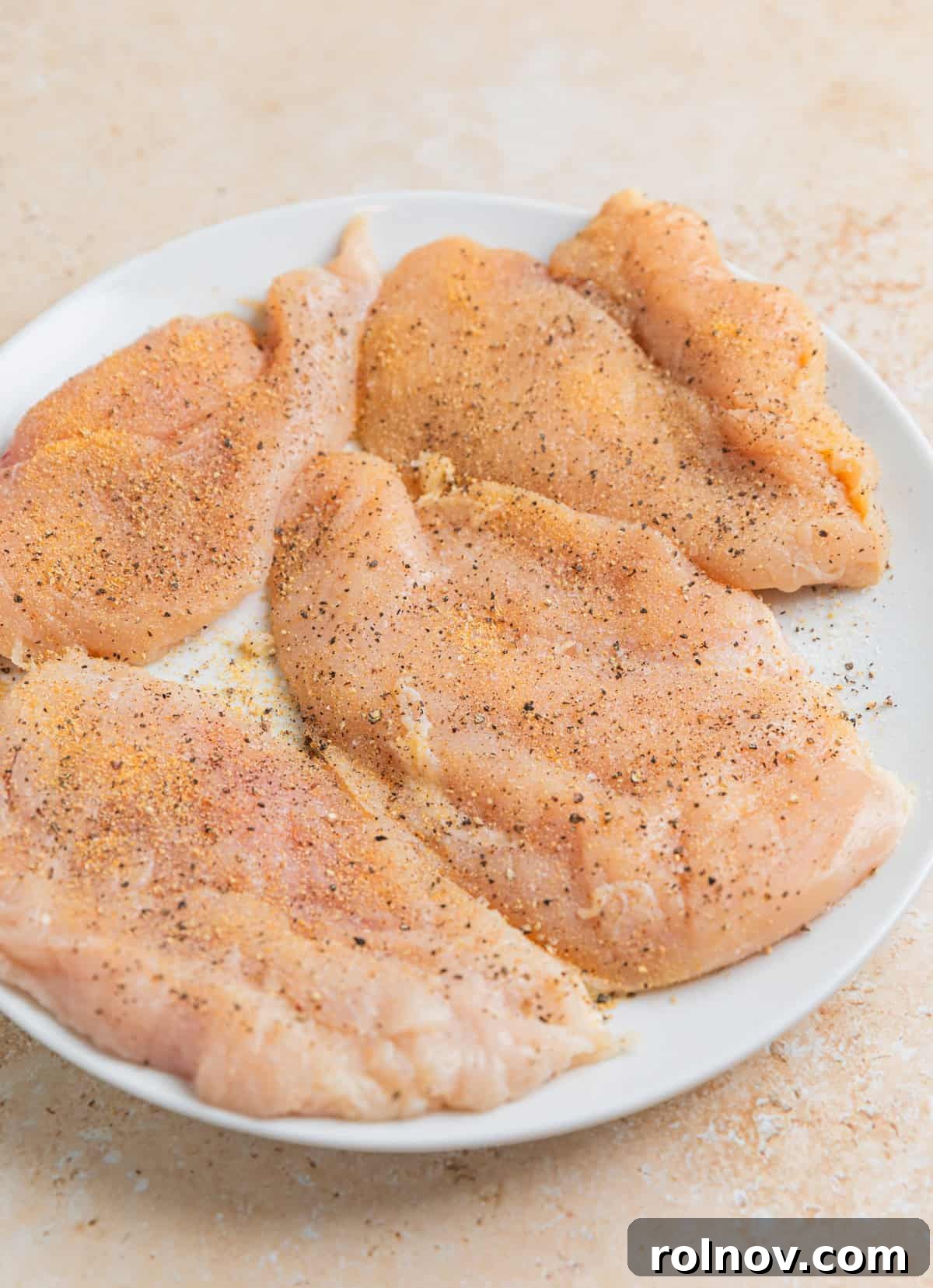 Seasoned thin chicken breasts on white plate before cooking.