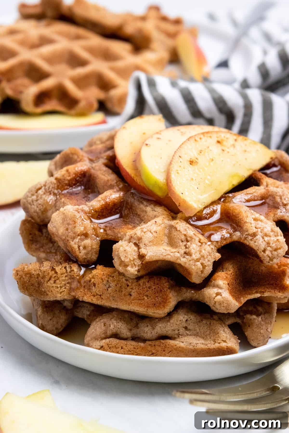 A beautiful stack of golden-brown vegan apple cinnamon waffles, topped with fresh apple slices and a light dusting of cinnamon.