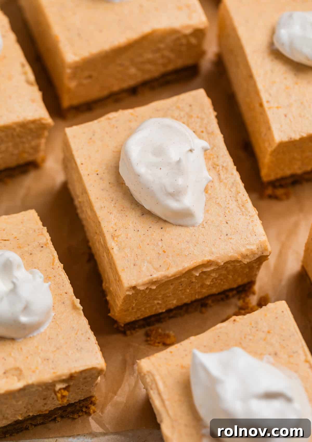 No bake pumpkin cheesecake bars on parchment topped with a dollop of whipped cream and a sprinkle of cinnamon.