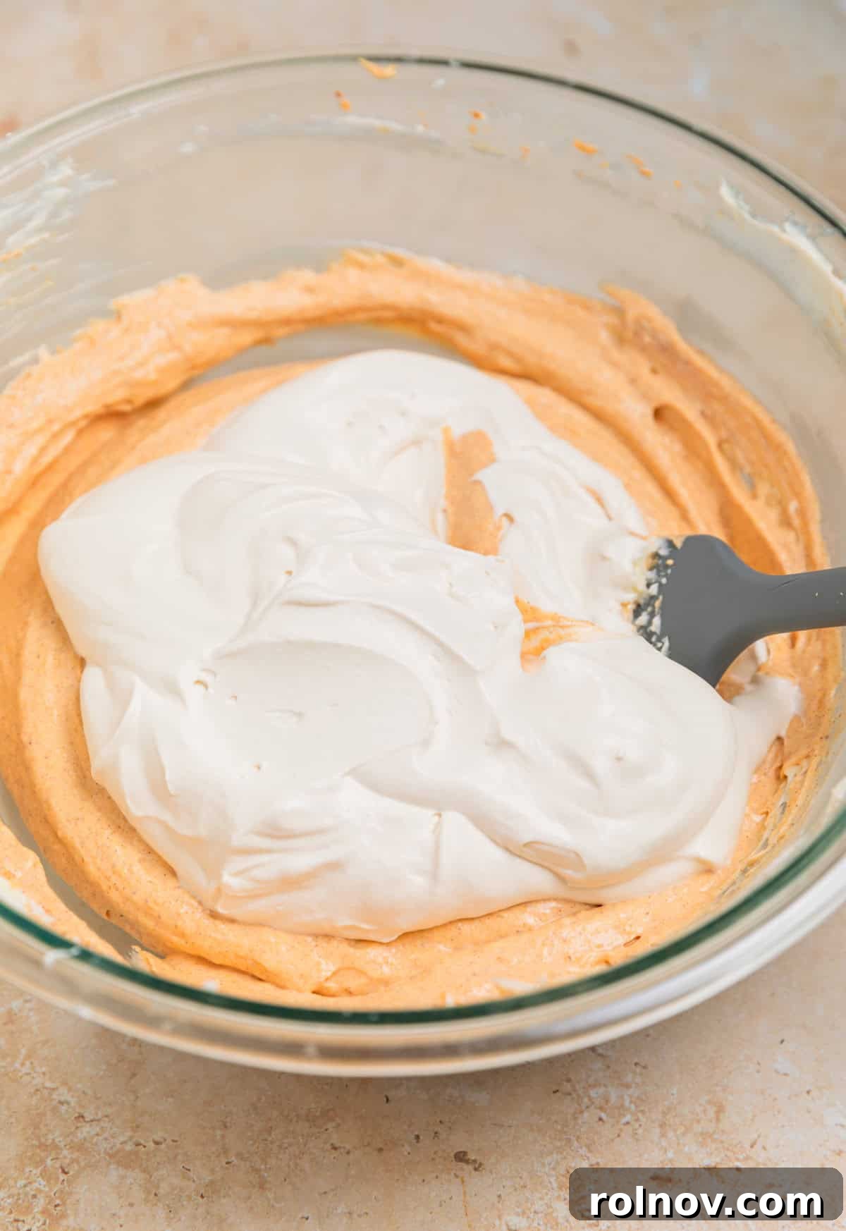 A portion of brown sugar whipped cream being gently added to the pumpkin cream cheese mixture in a bowl with a spatula, prior to folding.