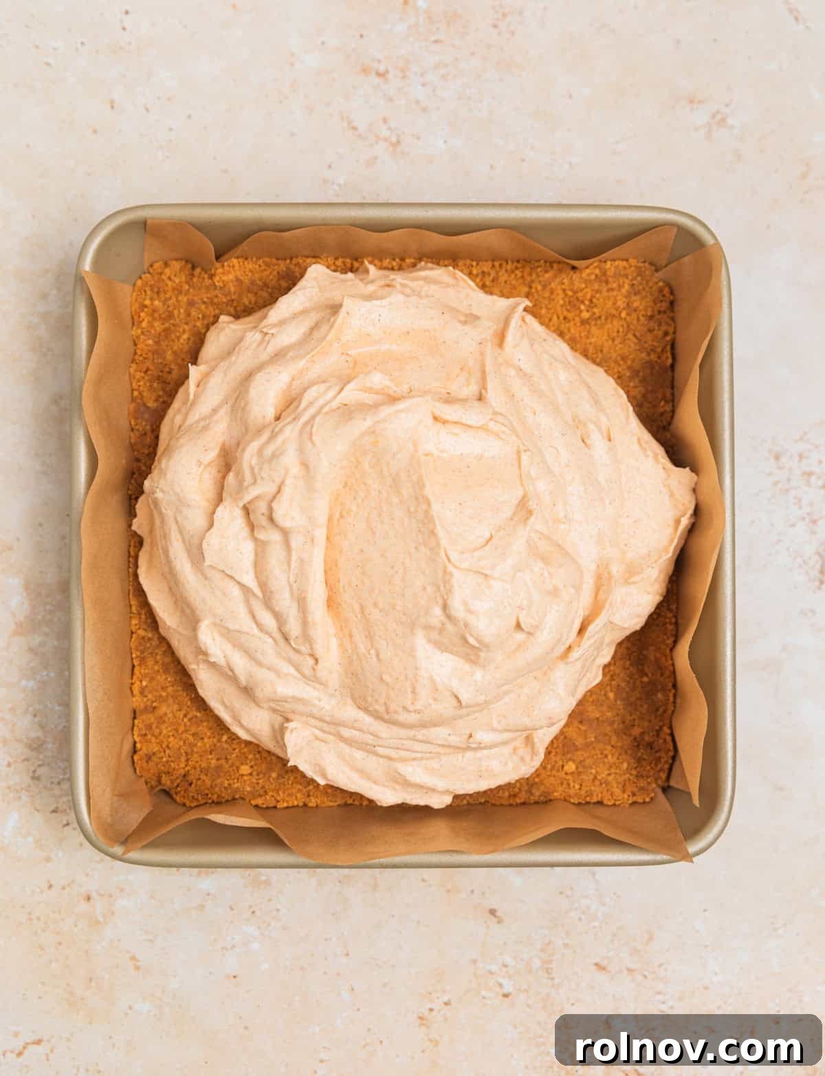 Pumpkin cheesecake filling being carefully spooned and spread over the ginger snap cookie crust in the square pan.
