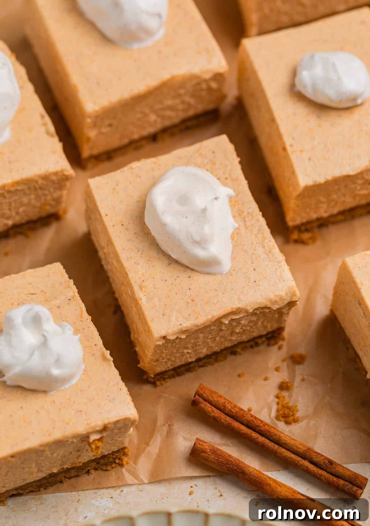 Slices of no bake pumpkin cheesecake arranged on parchment paper, each topped with a small dollop of whipped cream and a sprinkle of spice.