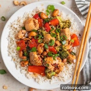 Baked cashew chicken over rice on plate with chopsticks, garnished with fresh herbs and crunchy cashews.
