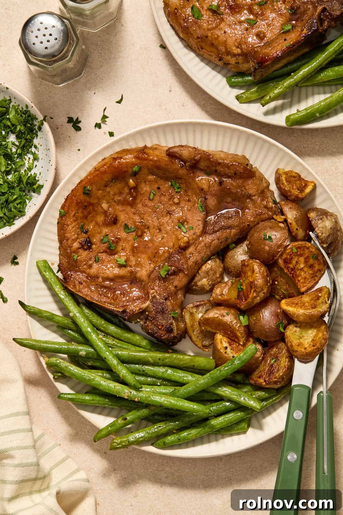 Quick & Flavorful Sheet Pan Pork Chops 11 A beautifully plated serving of honey garlic pork chops, accompanied by crispy parmesan potatoes and vibrant green beans, garnished with fresh herbs on a white plate.