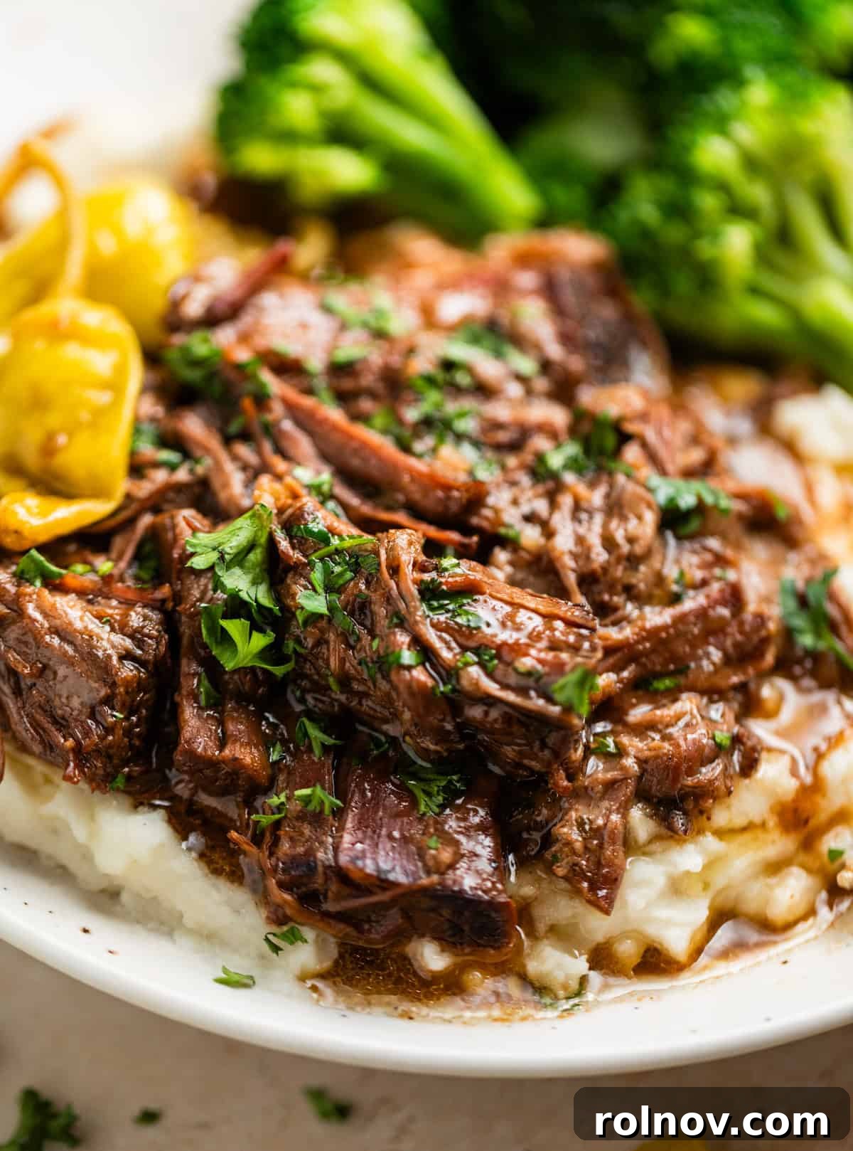 Melt-in-Your-Mouth Mississippi Pot Roast 2 Missisippi pot roast beef served over mashed potatoes with broccoli and pepperoncini peppers in the background.