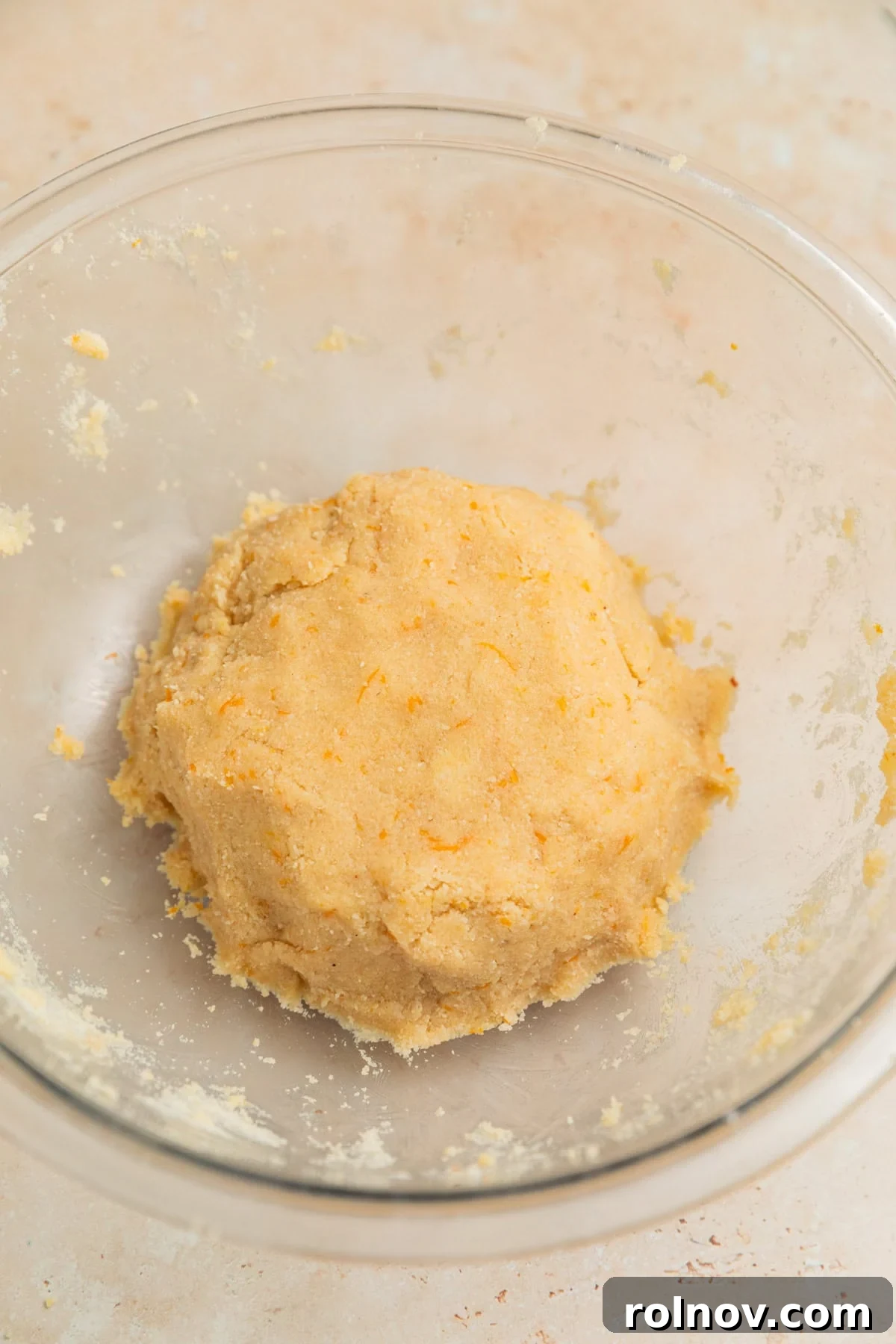 A perfectly formed ball of orange-infused almond flour cookie dough resting in a clear glass mixing bowl, showcasing its moist, uniform texture.