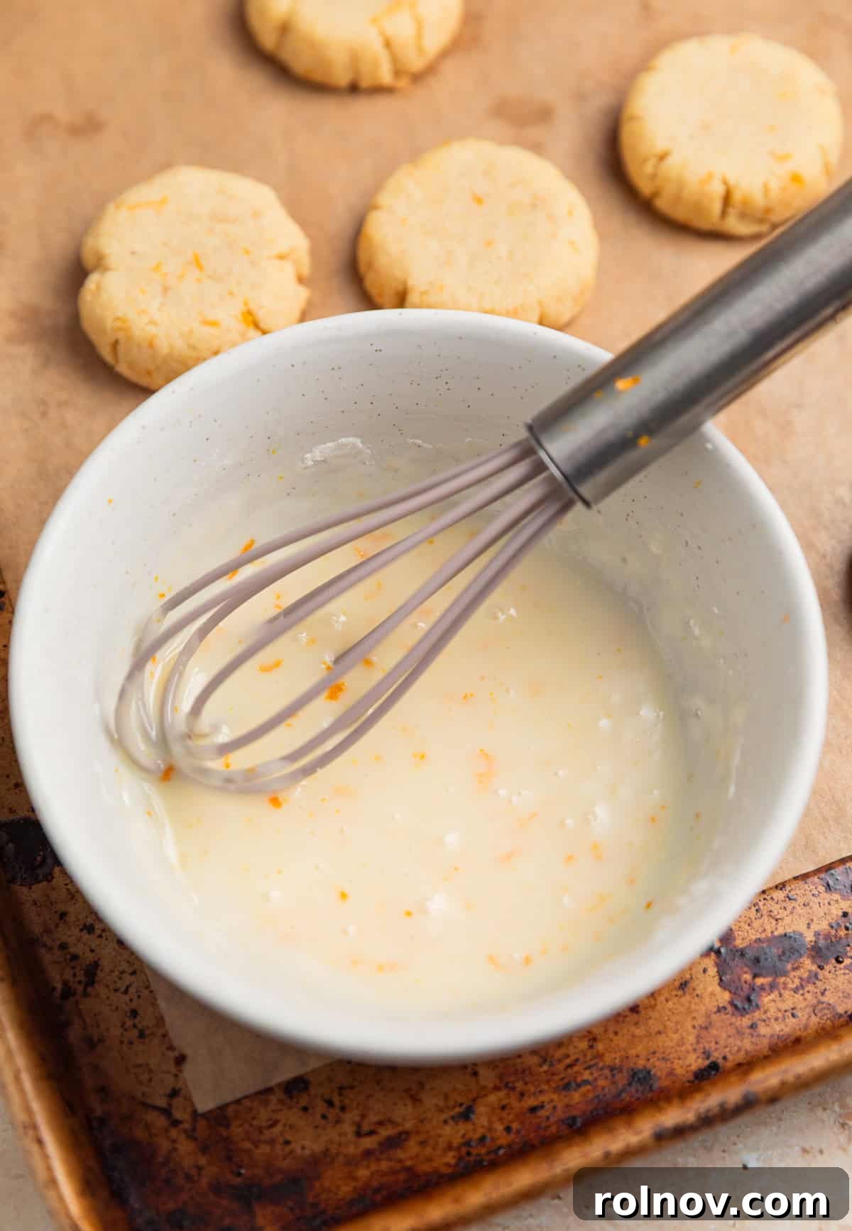 A bowl of glossy orange icing with a whisk resting in it, showcasing its smooth, vibrant consistency, ready for dipping.