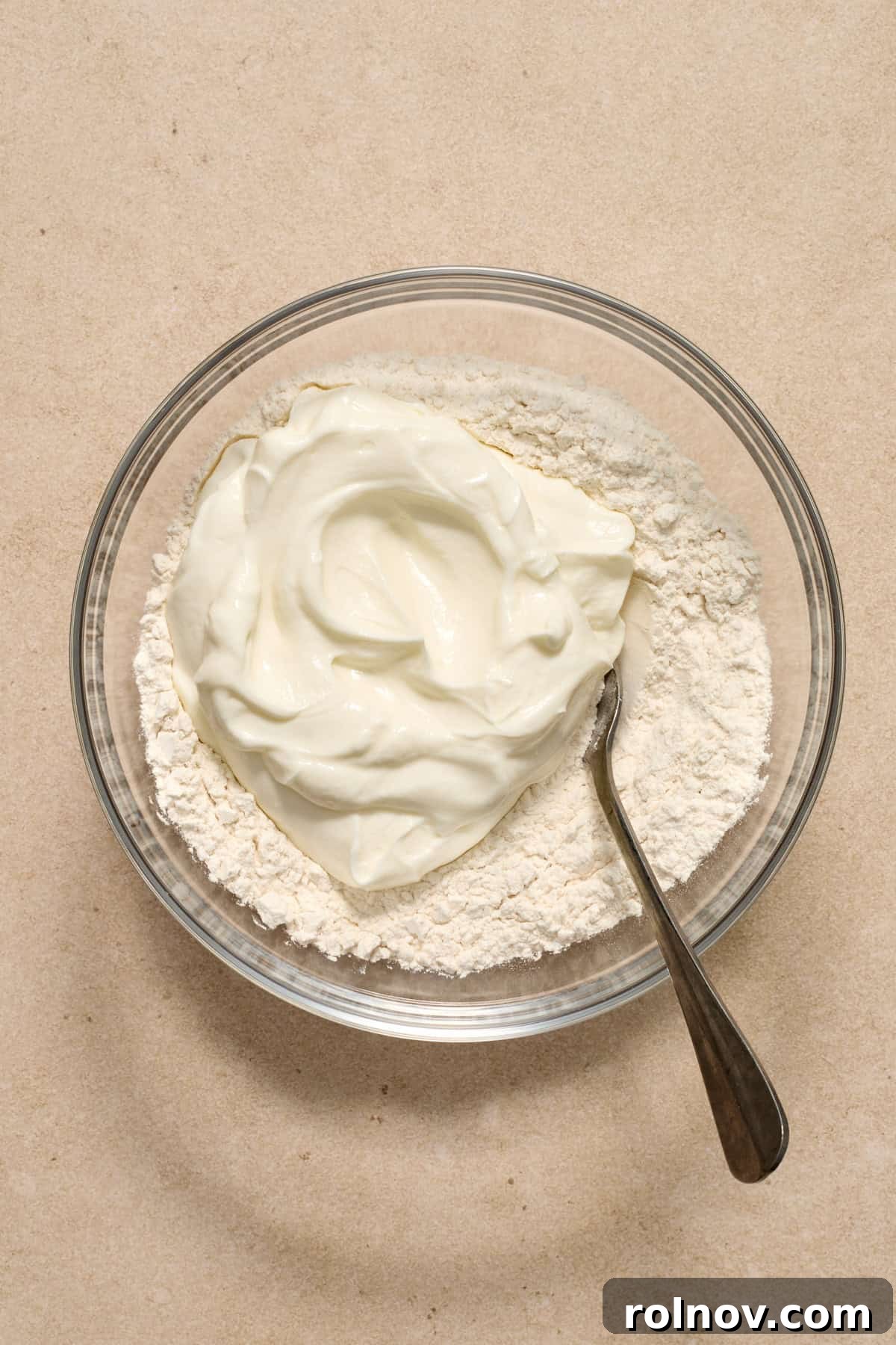 Adding Greek Yogurt to Flour Mixture Thick, plain Greek yogurt is carefully added to the bowl containing the dry flour mixture using a spoon.