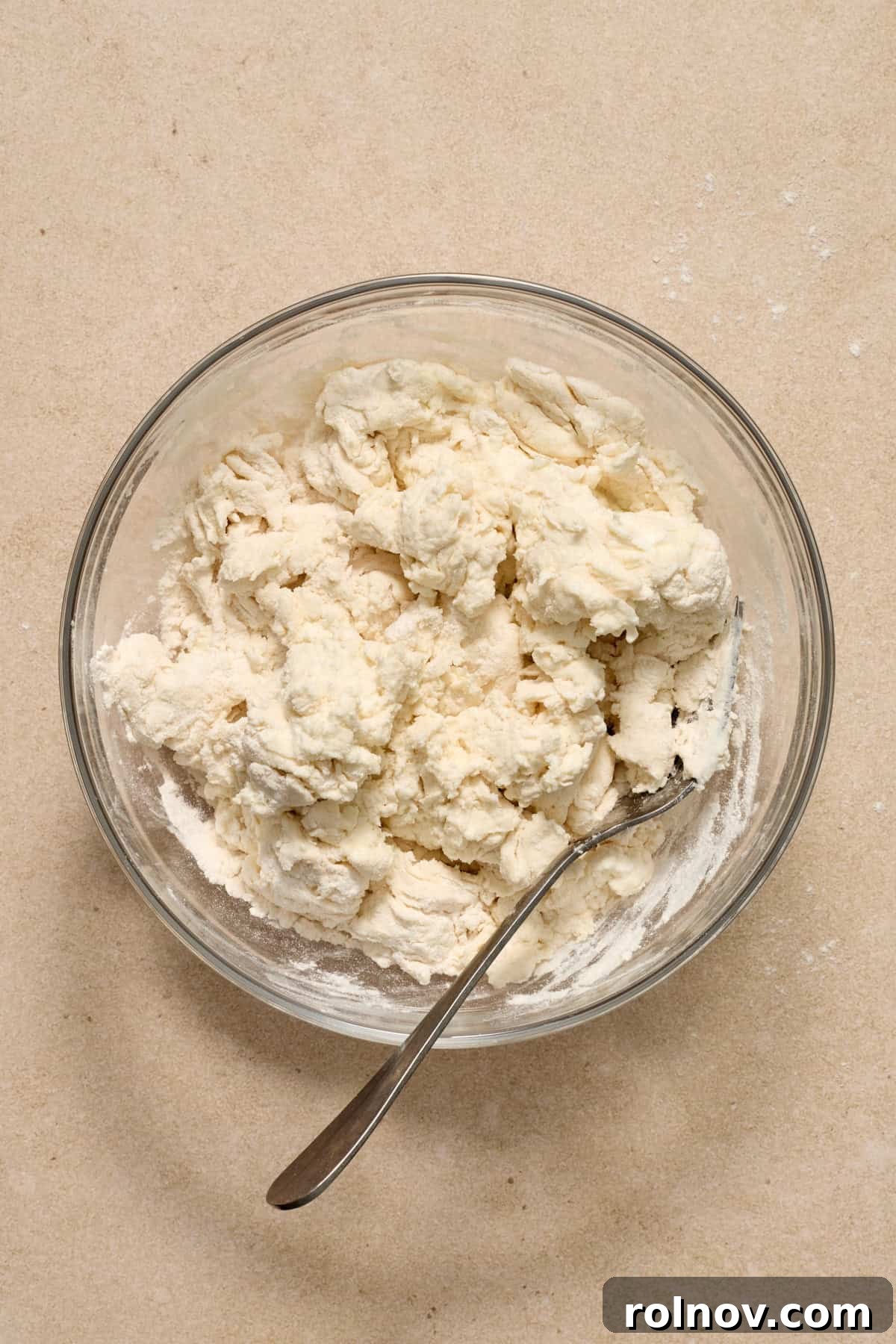 Mixing Yogurt into Bagel Dough The Greek yogurt bagel dough mixture in a glass bowl, partially combined with a metal spoon, showing its crumbly texture.