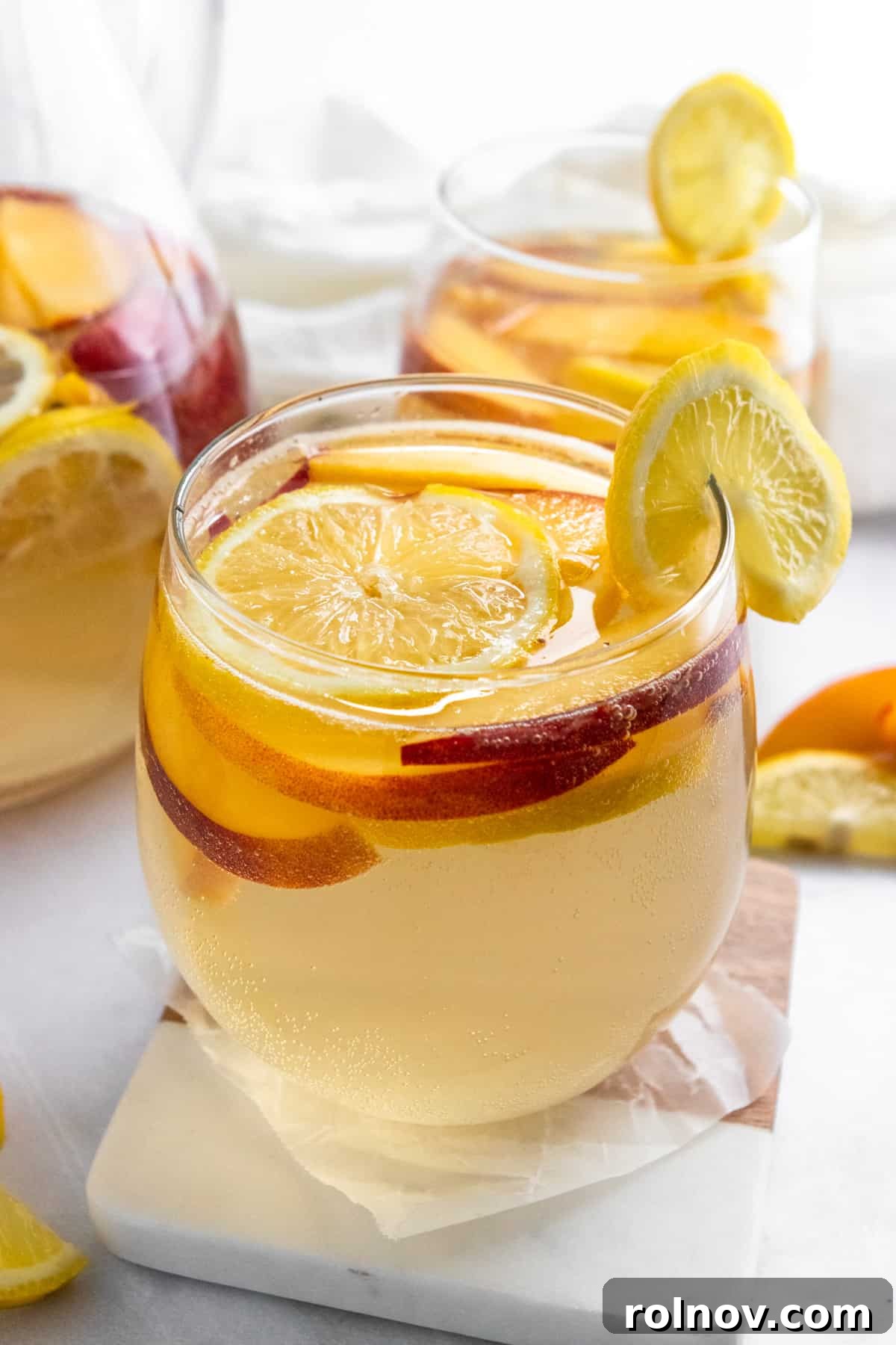 Sun-Kissed Peach Ginger Sangria 2 Close-up of a glass of Ginger Peach White Sangria, garnished with a lemon slice and fresh peach, sitting on a wooden coaster.