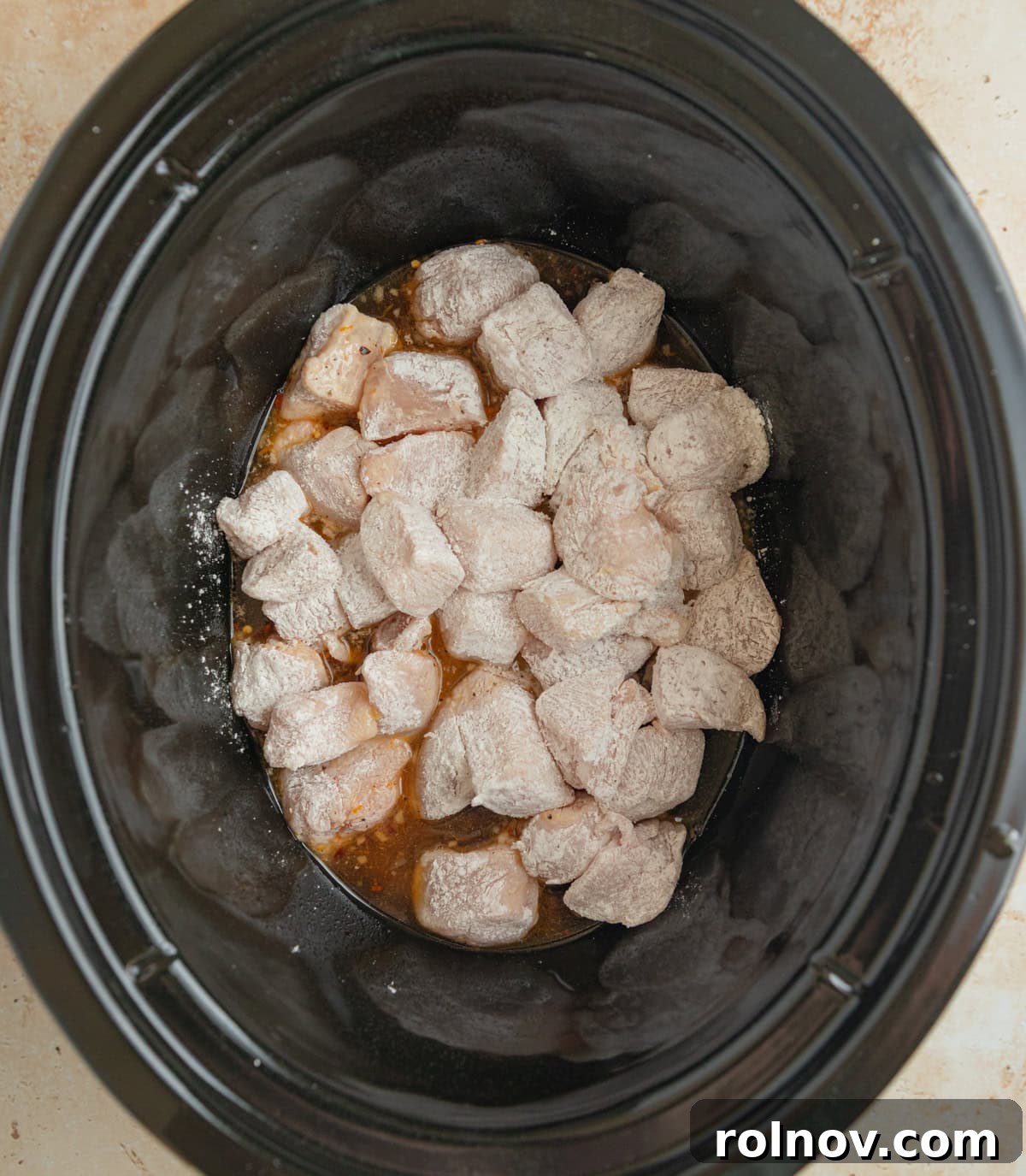 Zesty Slow Cooker Orange Chicken 7 The bottom of a slow cooker pot lightly coated with a thin layer of orange sauce, with cornstarch-coated chicken cubes placed over it.