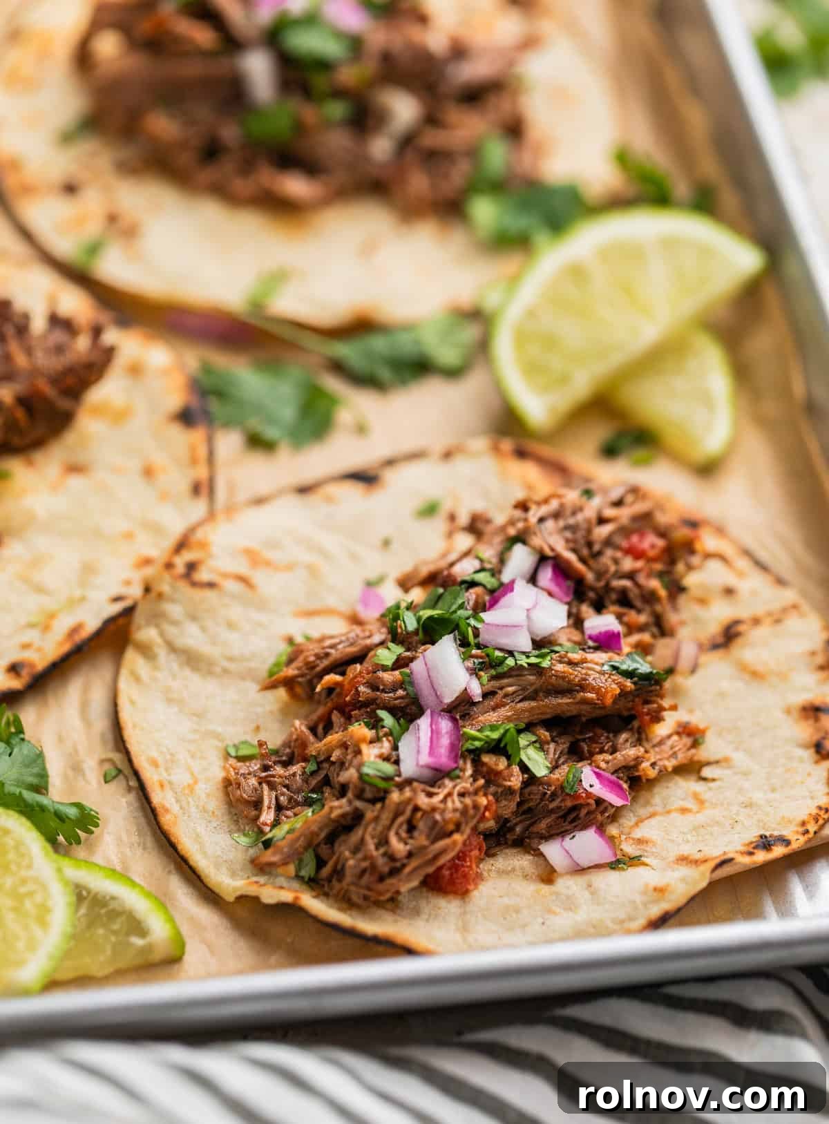Melt In Your Mouth Slow Cooker Beef Tacos 2 Shredded beef tacos on parchment lined pan topped with onion and cilantro.