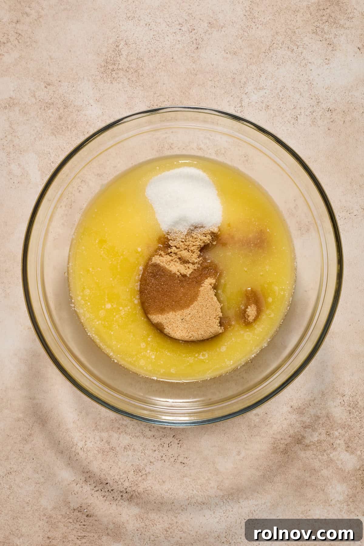 Melted butter, granulated sugar, and brown sugar combined in a clear glass mixing bowl, ready for mixing.