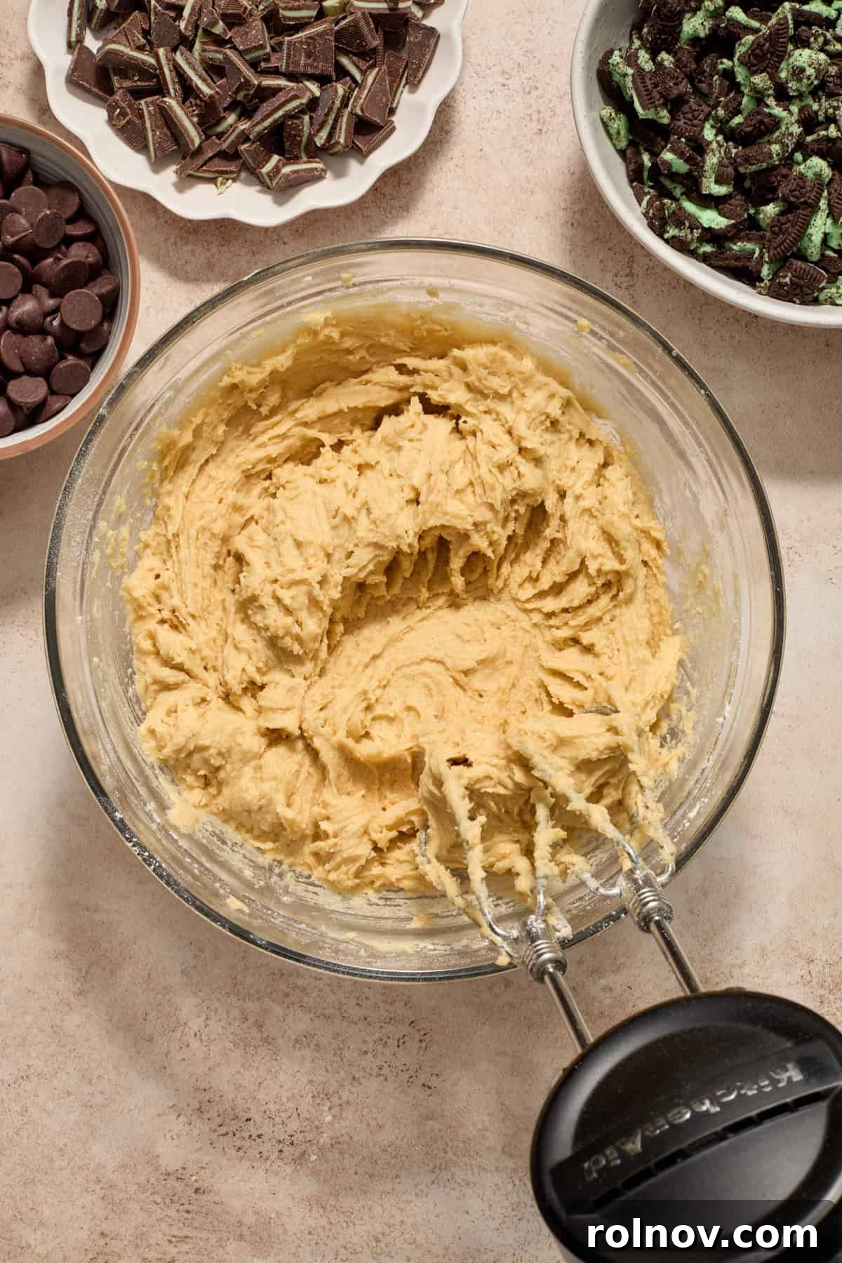 A bowl of mint chocolate cookie dough with a hand mixer, surrounded by smaller bowls containing chocolate chips, crushed mint Oreos, and chopped Andes mints as add-ins.