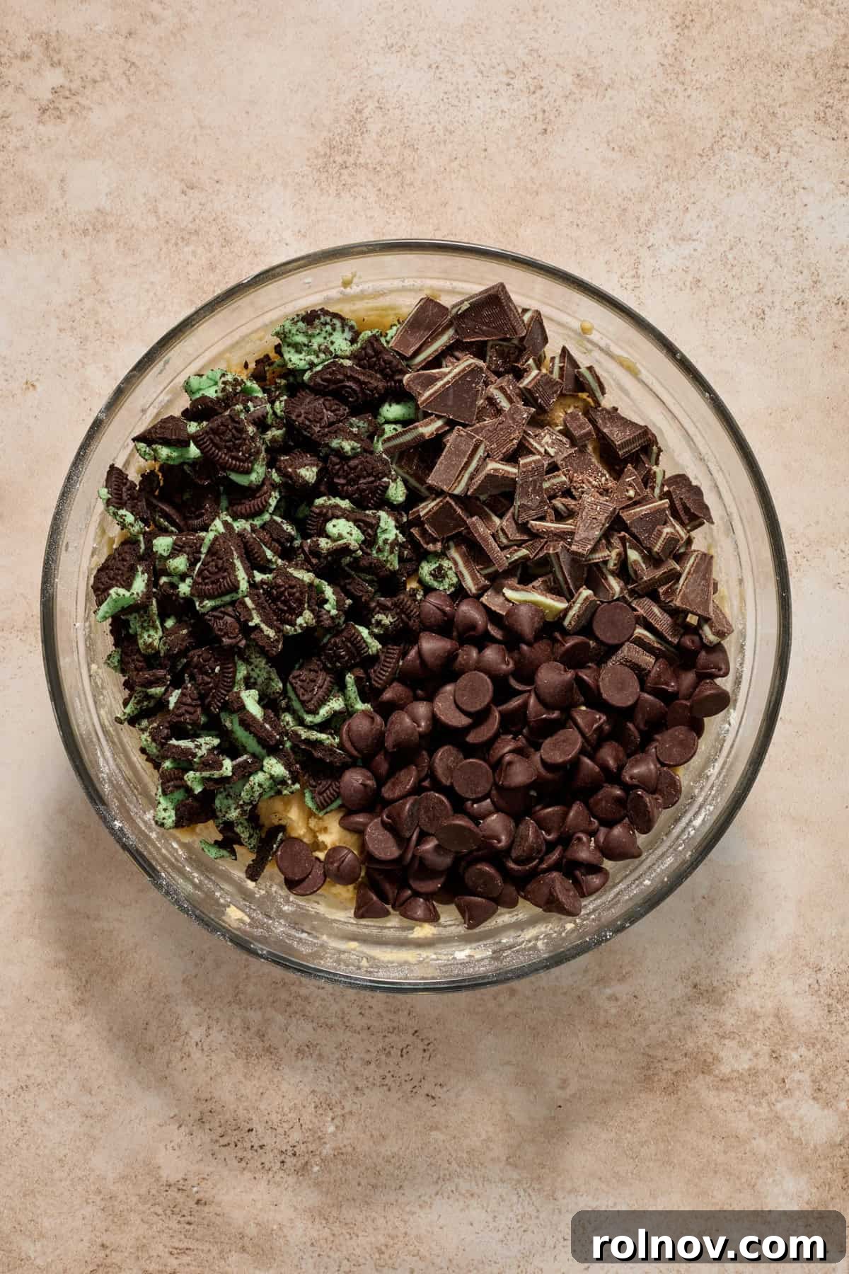 Close-up of cookie dough in a bowl, with a generous amount of dark chocolate chips, crushed mint Oreo cookies, and chopped Andes mints folded into it.