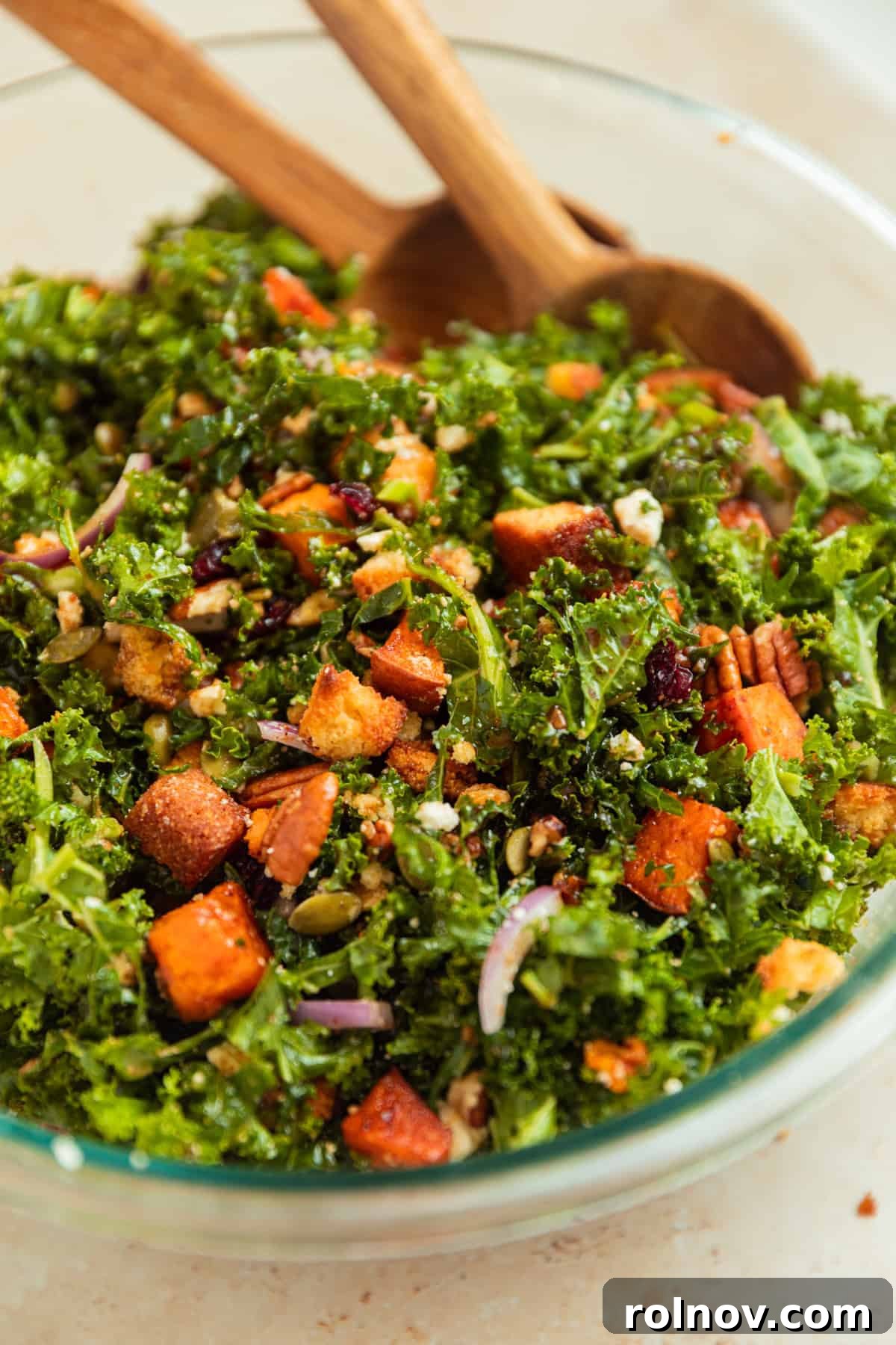 Roasted Sweet Potato Kale Salad 12 A close-up of balsamic dressing being drizzled over the tossed kale salad in a wooden bowl, with wooden spoons nearby for mixing.