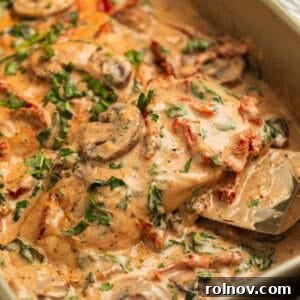 Creamy baked Tuscan chicken topped with mushrooms and parsley, being scooped from a pan.