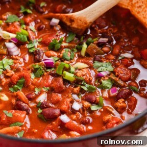 A vibrant red Dutch oven filled with ground turkey chili, topped with red onion, green onion, and cilantro, with a wooden spoon resting inside.