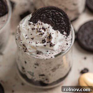 Individual Oreo cheesecake cups, generously topped with whipped cream and a whole Oreo cookie, surrounded by crumbs.