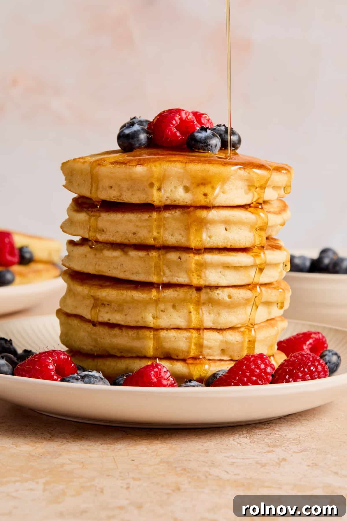 A tall stack of golden dairy-free pancakes generously drizzled with maple syrup and topped with fresh berries, sitting on a white plate.