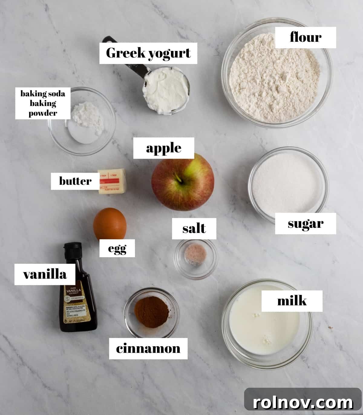 Neatly arranged baking ingredients on a kitchen counter: flour, sugar, cinnamon sticks, whole apple, and other baking essentials, ready for making apple donuts.