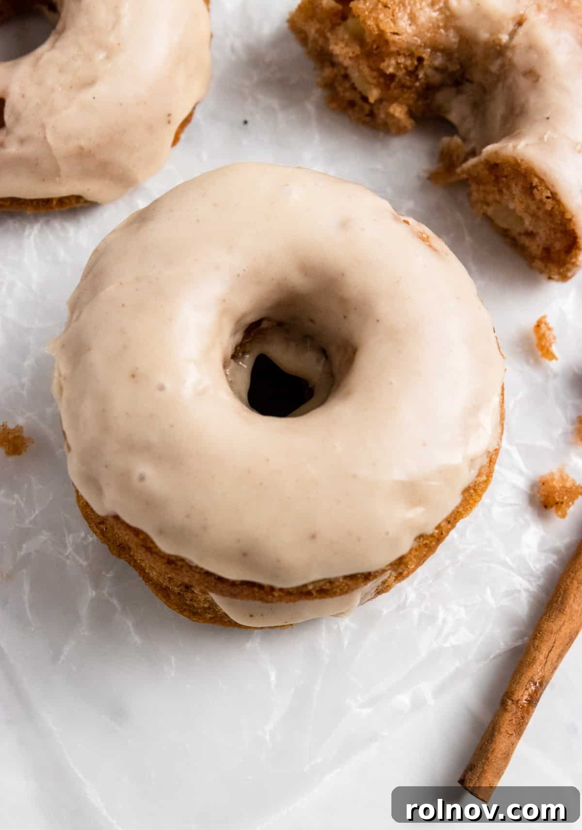 A close-up of a glazed apple cinnamon donut, half-eaten, revealing the soft interior and apple pieces, with a cinnamon stick garnish.