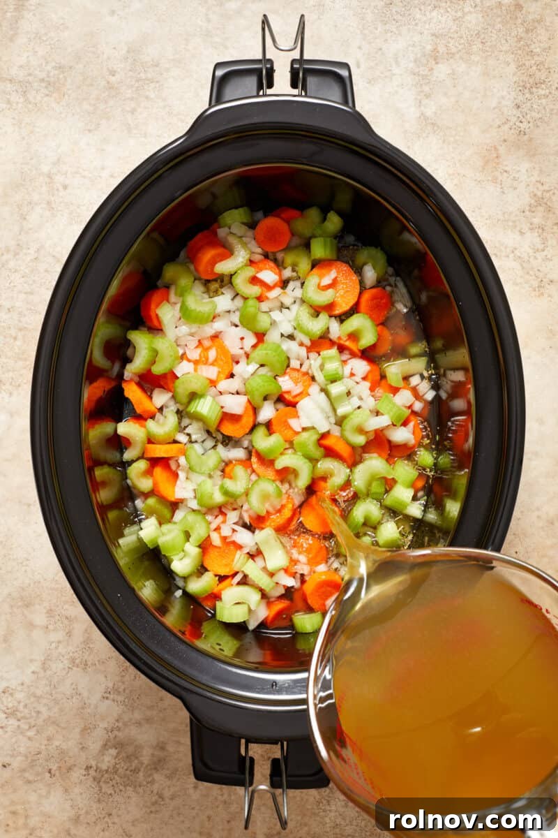 Celery, onion and carrots in slow cooker pot with broth being poured over.