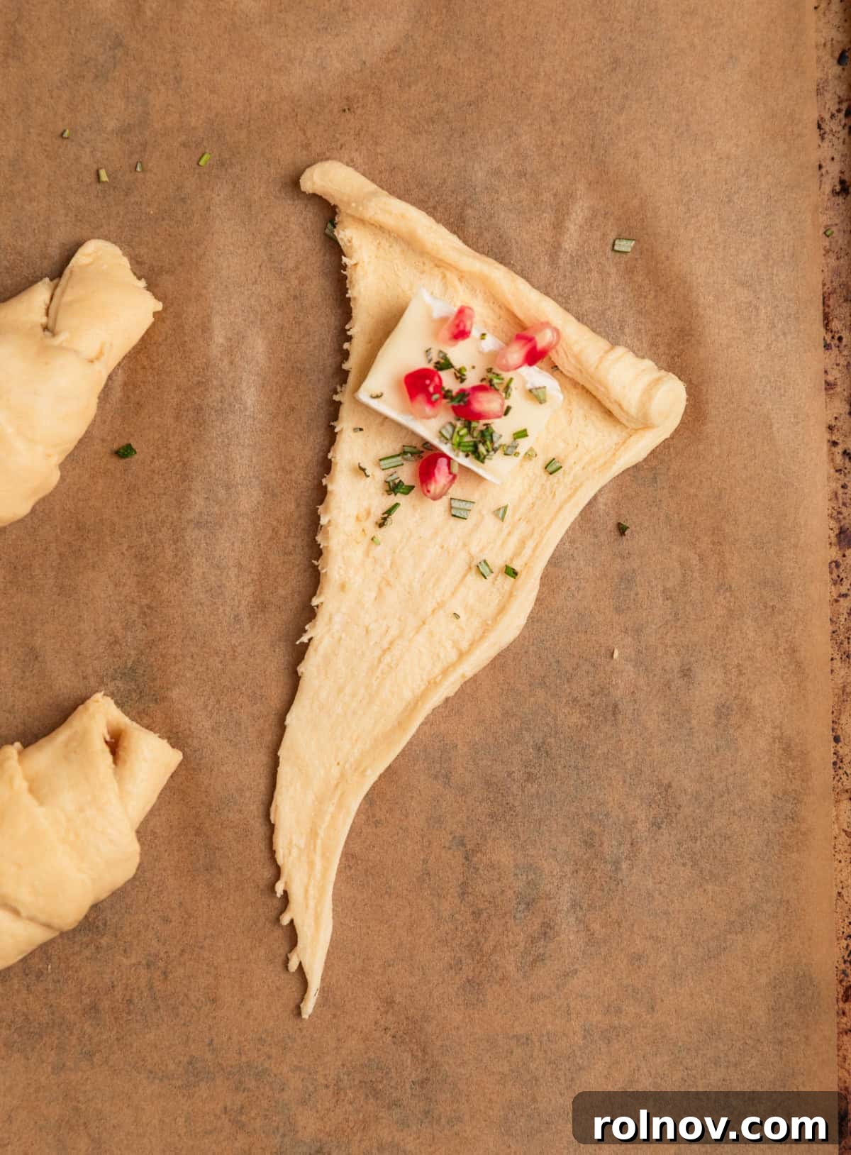 Golden Flaky Brie Bites 5 Crescent roll dough on parchment with brie, rosemary and pomegranate filling placed near the wide edge.