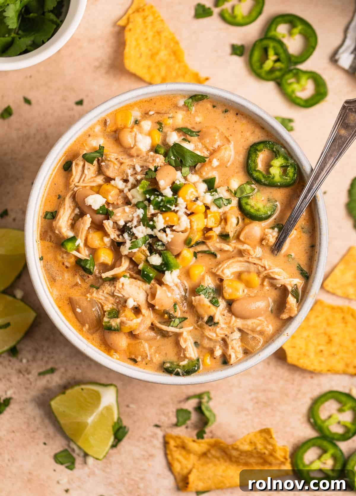 A bowl of creamy white chicken chili, garnished with fresh queso fresco, sliced jalapeños, and vibrant cilantro. Tortilla chips and lime wedges are artfully arranged alongside, inviting a spoonful into the rich soup.