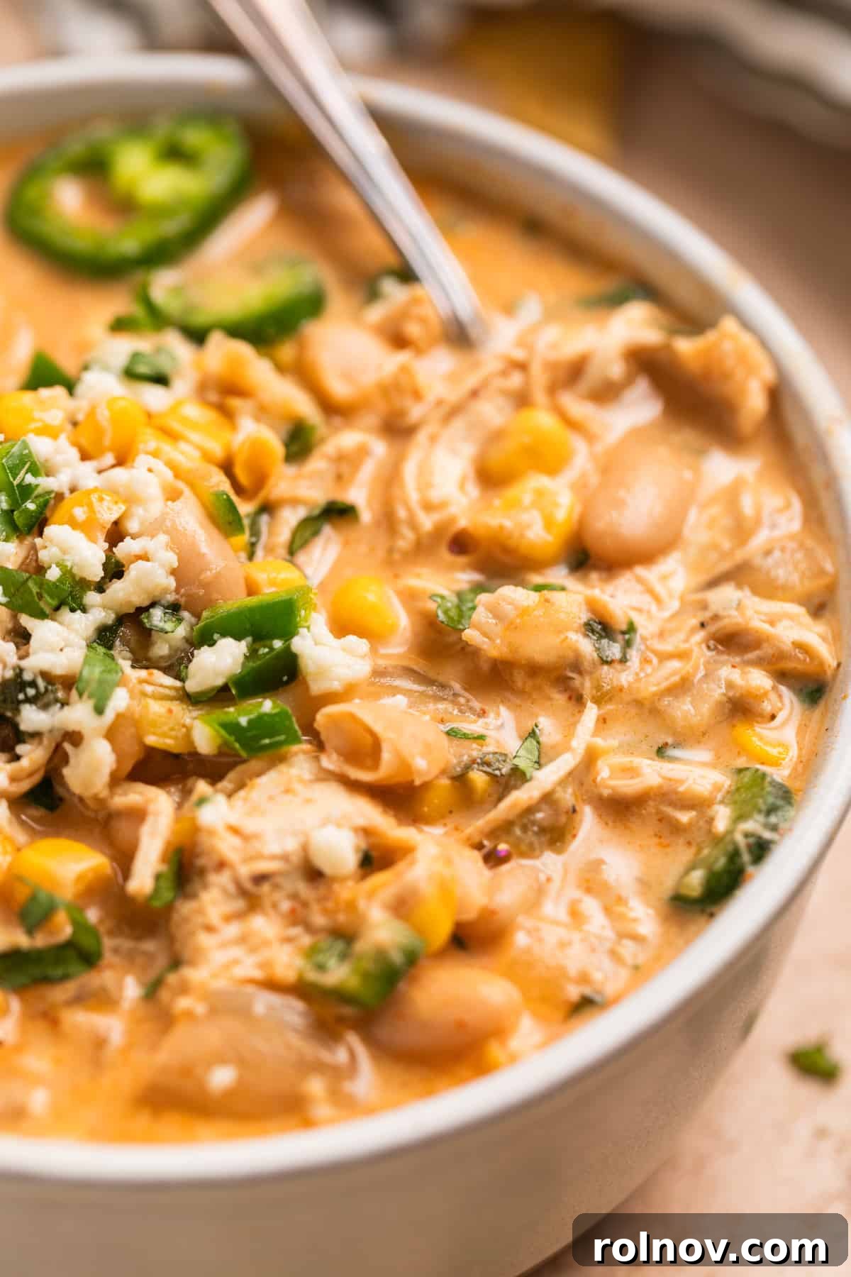 A close-up shot of a bowl of slow cooker white chicken chili, adorned with fresh corn, cilantro, shredded cheese, and sliced jalapeños, with a spoon ready for tasting.