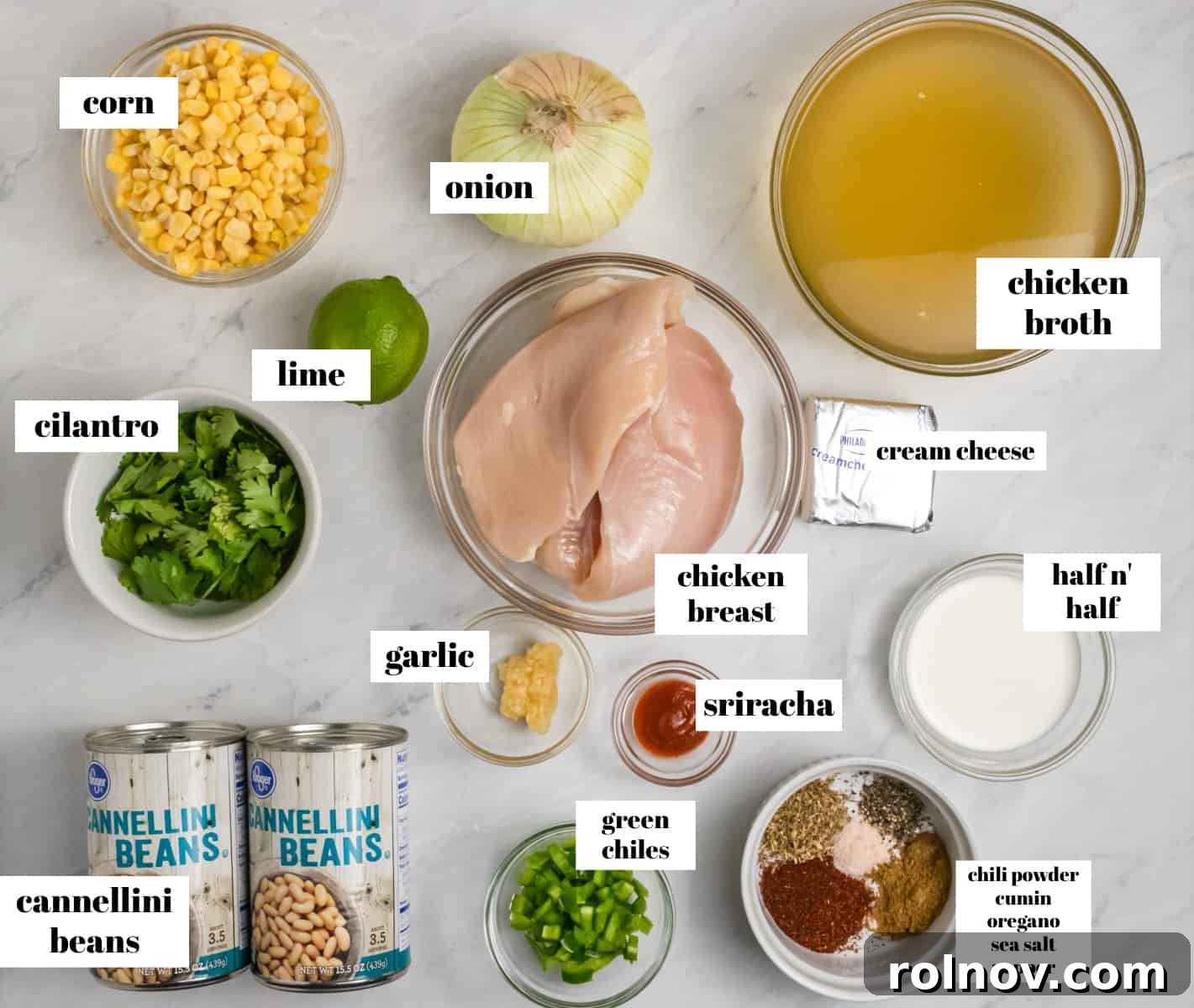 Individual ingredients for slow cooker creamy white chicken chili laid out on a kitchen counter, labeled: chicken, cilantro, cream cheese, broth, beans, corn, spices.