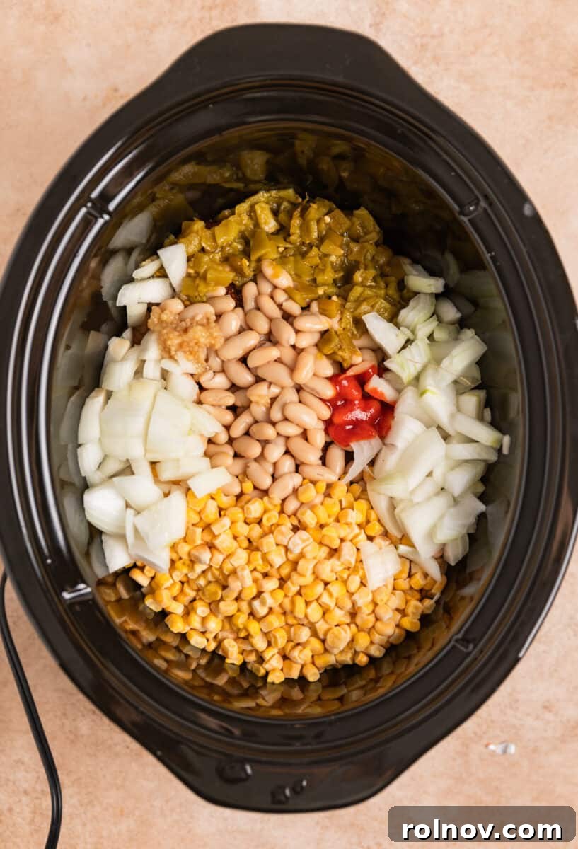 A slow cooker filled with chicken, corn, onion, green chiles, garlic, and a drizzle of sriracha, layered over the seasoned chicken.