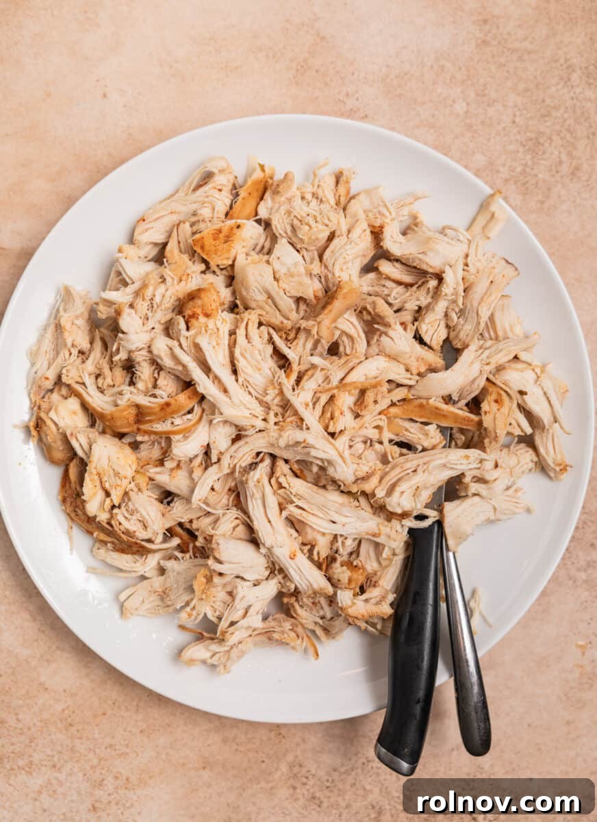 Cooked chicken breast shredded into tender pieces on a plate, with a knife and fork nearby.