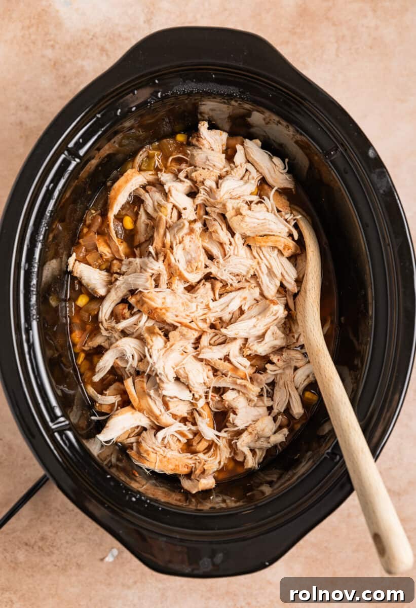 Shredded chicken being stirred back into the simmering chili in the slow cooker with a wooden spoon.