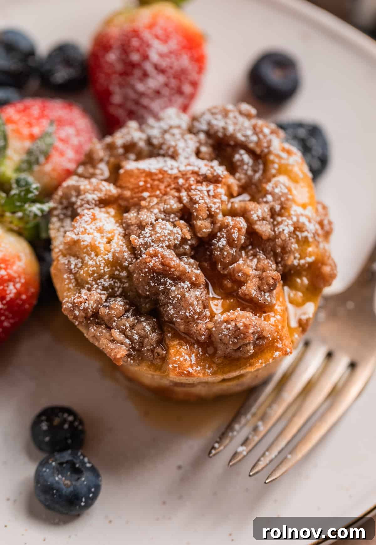 Golden French Toast Bites 3 French toast muffin with streusel topping, maple syrup and powdered sugar on plate with berries and fork.