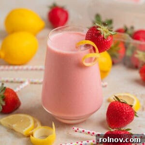 Wine glass with strawberry lemon smoothie with lemon zest and strawberry garnish and berries, lemon slices and straws surrounding.