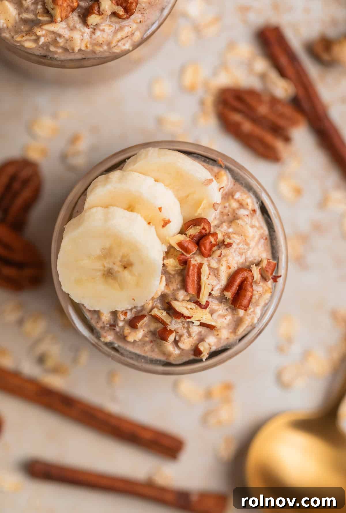 Glass jar filled with banana bread overnight oatmeal, elegantly garnished with banana slices, chopped pecans, and cinnamon sticks. Surrounding the jar are loose pecans and a spoon, highlighting its inviting presentation.
