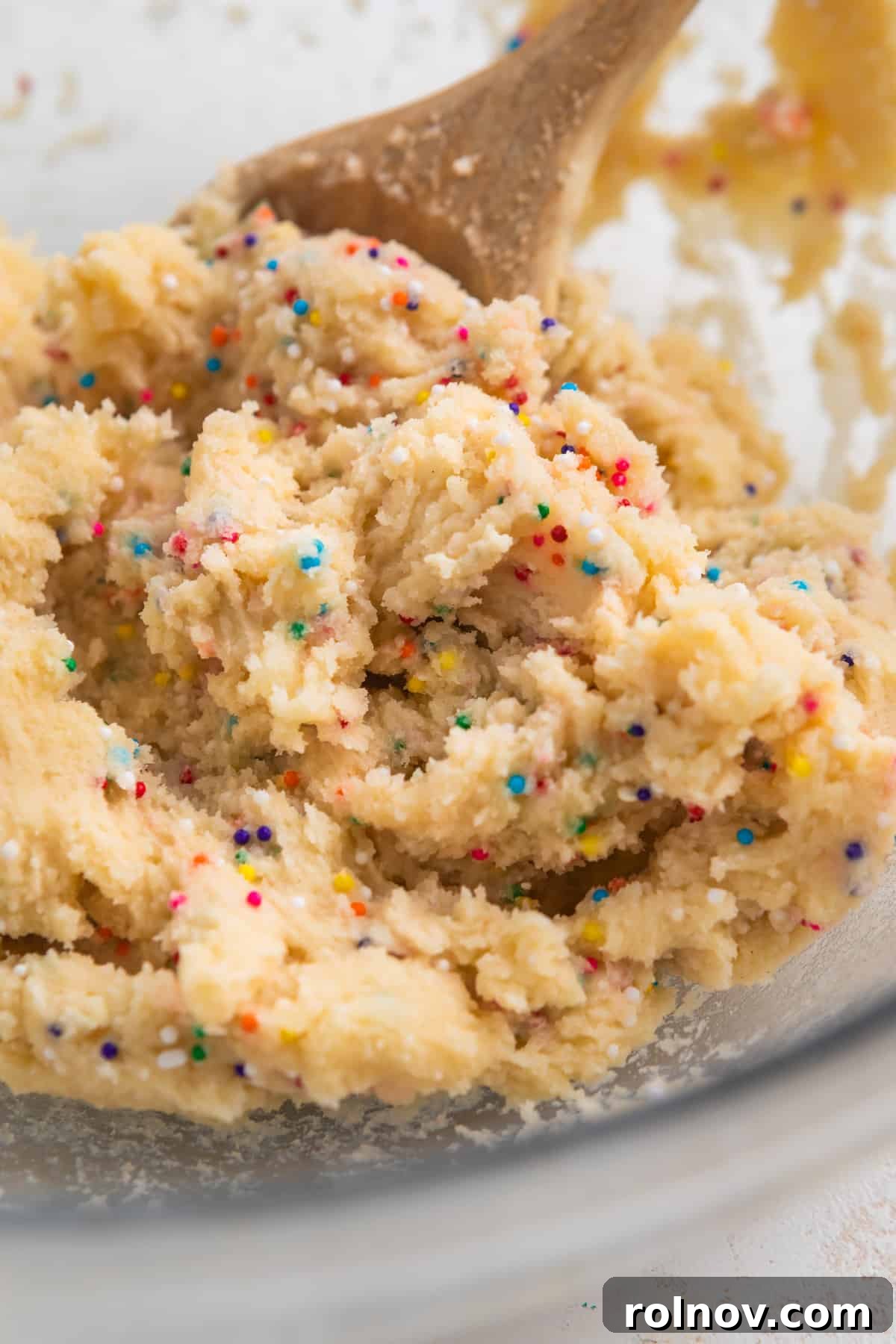 Spoonable Sugar Cookie Dough 5 A mixing bowl filled with freshly prepared edible sugar cookie dough, adorned with colorful sprinkles, with a wooden spoon poised for sampling.