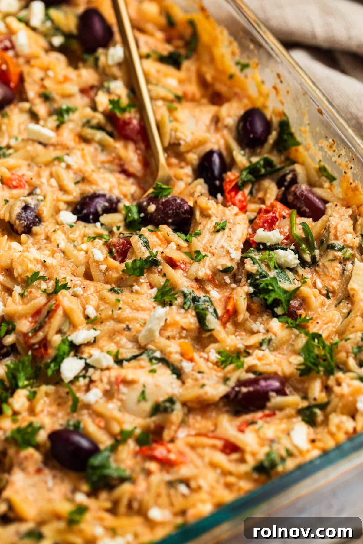 Aegean Chicken and Orzo Bake 2 A glass baking dish filled with cooked Greek chicken and orzo, with a spoon scooping out a portion, highlighting the creamy melted feta, tender chicken, and burst cherry tomatoes.