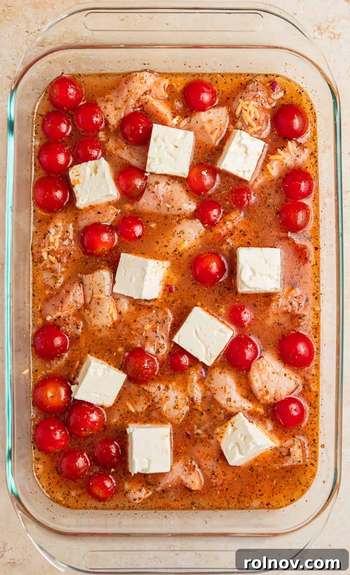 Aegean Chicken and Orzo Bake 6 Uncooked orzo pasta, seasoned chicken pieces, whole cherry tomatoes, and large cubes of feta cheese arranged in a baking pan, with chicken broth and lemon juice added, ready for mixing and baking.